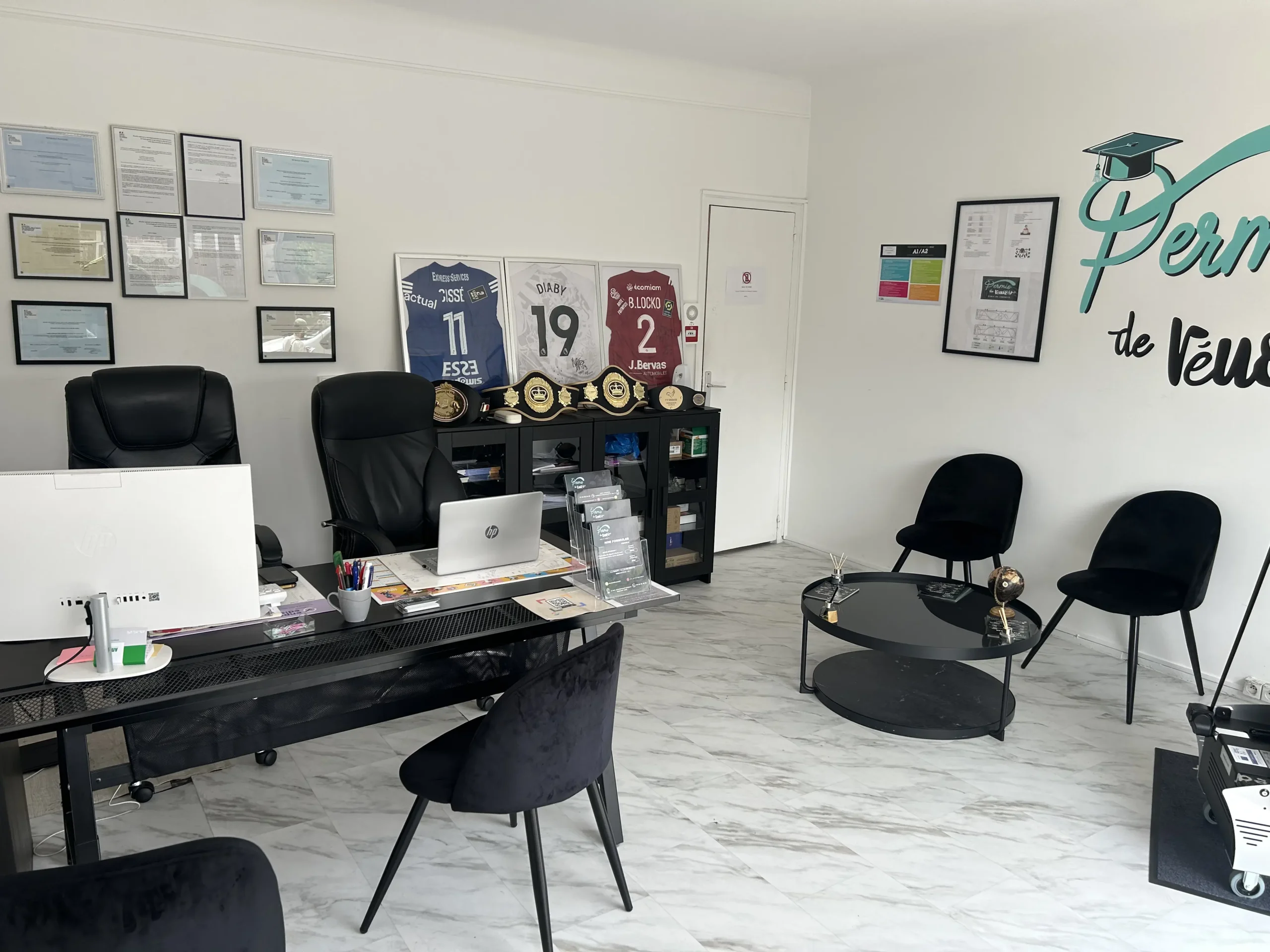 auto-ecole-villejuif-bureau-permis-de-reussir Intérieur du bureau de l’auto-école Permis de Réussir à Villejuif avec deux postes de travail, diplômes encadrés, maillots de football, et trophées exposés.